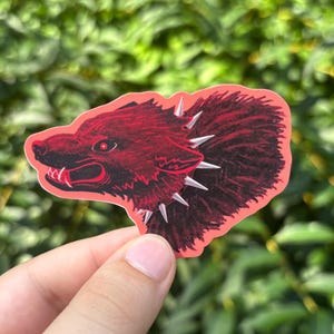 May include: A red sticker featuring a wolf's head with a spiked collar. The wolf has a menacing expression with visible fangs and a glowing red eye. The sticker is outlined in red and has a textured appearance.