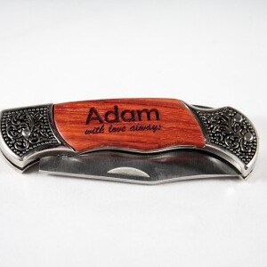 May include: A personalized pocket knife with a wooden handle engraved with the name "Adam" and the phrase "with love always." The knife features a silver-colored metal frame with ornate floral detailing.