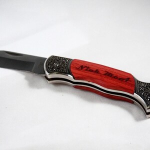 May include: A folding pocket knife with a black blade and a red wooden handle. The handle has silver accents and decorative metalwork. The text "Nick Wood" is engraved on the handle.