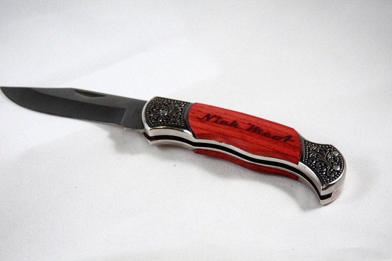 May include: A folding pocket knife with a black blade and a red wooden handle. The handle has silver accents and decorative metalwork. The text "Nick Wood" is engraved on the handle.