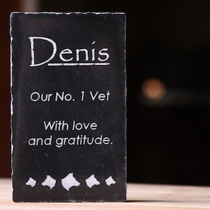 May include: Black slate plaque with white lettering that reads "Denis, Our No. 1 Vet, With love and gratitude."  The plaque has five white paw prints at the bottom.