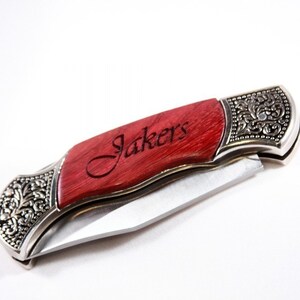 May include: A folding pocket knife with a red wooden handle and silver-colored metal accents. The handle is engraved with the word "Jakers". The knife blade is silver and partially open, showcasing the sharp edge. The metal accents feature an ornate floral design.
