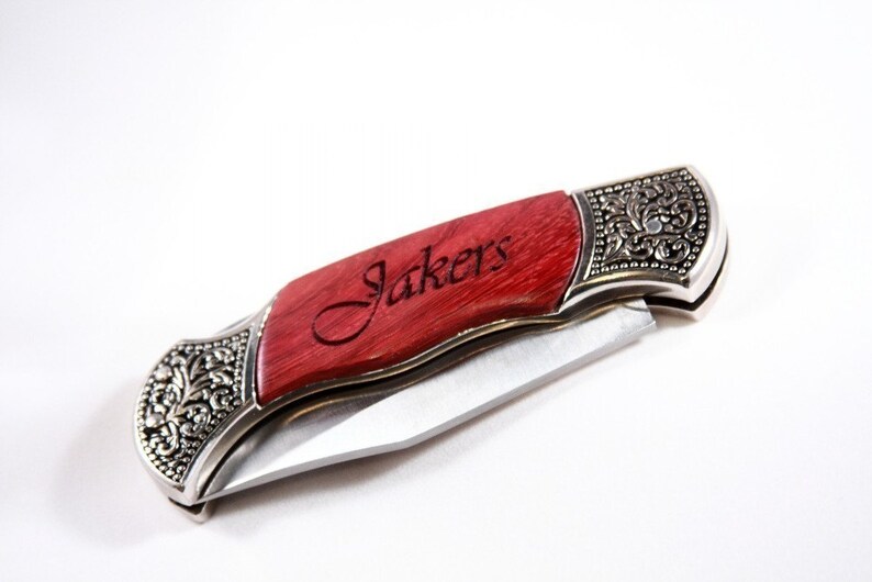 May include: A folding pocket knife with a red wooden handle and silver-colored metal accents. The handle is engraved with the word "Jakers". The knife blade is silver and partially open, showcasing the sharp edge. The metal accents feature an ornate floral design.