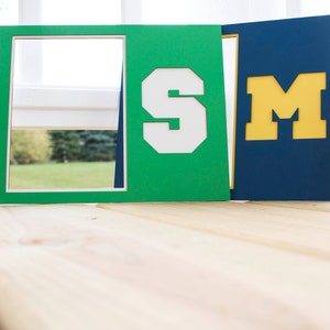 Peut inclure: Deux cadres décoratifs, l'un vert avec un "S" blanc et l'autre bleu marine avec un "M" jaune. Les cadres sont en papier épais ou carton et sont conçus pour contenir une photo ou une œuvre d'art. Les cadres sont exposés sur une surface en bois clair.