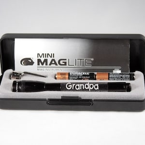 Puede incluir: Una linterna negra Mini Maglite con la palabra "Grandpa" grabada en ella. La linterna está en una caja negra con una etiqueta blanca que dice "Mini Maglite" y "Mag Instrument".