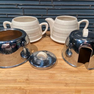 May include: A cream ceramic creamer and sugar bowl set with horizontal stripe detailing. The set includes a chrome-plated sugar bowl with a lid and a chrome-plated creamer holder. The items are arranged on a wooden surface.