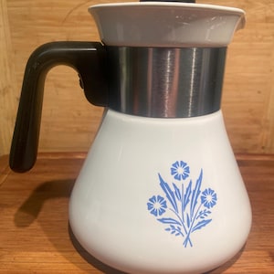 May include: A white ceramic teapot featuring a blue floral pattern on the front. The teapot has a wide base, a brushed metal band around the upper section, a black handle, and a black lid. The design includes three stylised blue flowers.