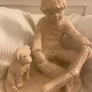 Könnte beinhalten: Hellbeige Skulptur eines Kindes, das mit einem Welpen sitzt. Das Kind hält einen langen Gegenstand. Der Welpe sitzt neben dem Kind. Die Skulptur steht auf einem runden Sockel.