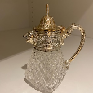 Könnte beinhalten: Eine Vintage-Sahnekännchen mit einem transparenten, rautenförmigen Glaskörper und verzierten silberfarbenen Metallakzenten. Der Deckel und der Griff sind mit floralen Designs und einem Löwenkopf verziert. Das Sahnekännchen hat eine goldfarbene Oberseite und einen gebogenen Griff.