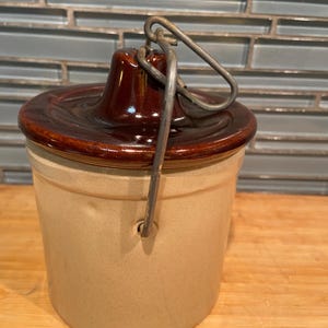 May include: A vintage stoneware crock with a brown lid and handle. The cylindrical container is beige with a dark brown lid and a metal handle. The lid has a raised center. The crock is likely used for food storage.