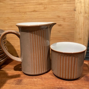 Può includere: Un set di brocca per la panna e zuccheriera in ceramica marrone con linee verticali. La brocca ha un manico e un beccuccio. La zuccheriera è rotonda.