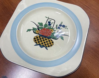 May include: A square, cream-colored plate with a blue rim. The center of the plate features a floral design with a basket, flowers, and leaves in shades of red, yellow, blue, and green.