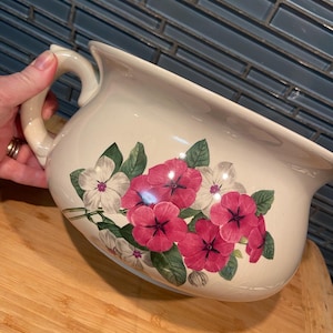 Pode incluir: Um bacio de cerâmica de cor creme com uma pega lateral e um padrão floral. O design apresenta cachos de flores cor-de-rosa vibrantes e brancas com folhas verdes pintadas na superfície frontal do recipiente arredondado de bordo largo.