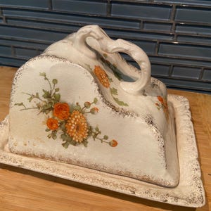 May include: A cream-coloured ceramic cheese dish with a domed cover and a rectangular base. The piece features a hand-painted floral design with orange chrysanthemums and green foliage, accented by a distressed brown speckled edge finish.