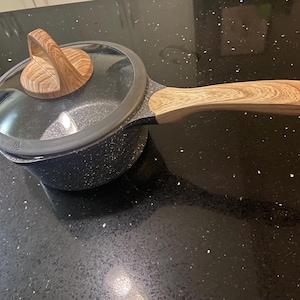 Puede incluir: Una cacerola negra y gris moteada con un mango de madera y una tapa de vidrio. La tapa tiene un pomo de madera.