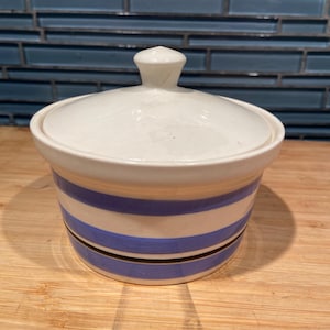 May include: A white ceramic lidded jar with blue and black horizontal stripes. The jar sits on a wooden surface with a blue tile background. The lid has a small knob on top.