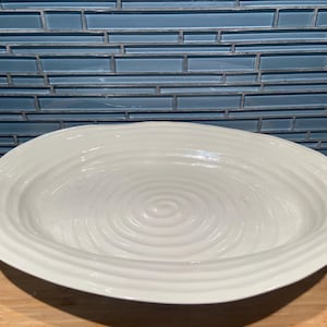 May include: An off-white ceramic serving bowl, oval in shape. The bowl features a rippled interior design and a raised rim. It sits on a wooden surface, with a blue tiled backsplash in the background. The bowl is approximately 35 cm in length.