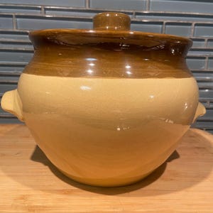 May include: A two-tone ceramic pot with a lid. The pot is light beige at the base, transitioning to a darker brown at the top, with a matching brown lid. It features two small handles on the sides.
