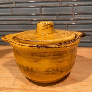 May include: A round, yellow ceramic lidded bowl with two side handles. The bowl features a glossy finish with dark brown, distressed-style brush strokes along the rim, the lid, and the base, creating a textured, weathered appearance.