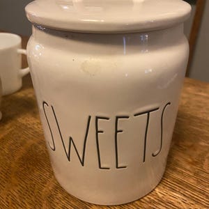 May include: A white ceramic jar with a lid and handle, labelled "SWEETS" in black lettering. The jar is cylindrical with a slightly tapered shape and sits on a wooden surface. The lid has a small handle for easy removal.
