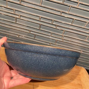 May include: A speckled blue ceramic mixing bowl with a brown rim. The bowl is held by a hand and sits on a wooden surface. The background features a blue and grey tiled wall.