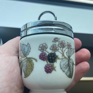 Pode incluir: Um cozedor de ovos de porcelana branca com tampa de metal cromado polido e alça circular. O corpo apresenta uma ilustração botânica de amoras, incluindo bagas roxas escuras e pretas, folhas verdes e pequenas flores brancas.