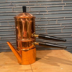 May include: A copper espresso maker with black handles and a spout. The coffee pot is made of shiny copper and has a black handle on the top. The espresso maker is on a wooden surface.