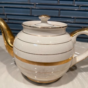 May include: A white ceramic teapot with gold accents. The teapot features a gold handle, spout, and lid finial. Gold stripes encircle the body of the teapot. The teapot is sitting on a white plate.