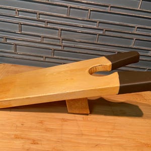 May include: A wooden boot jack with a light tan natural wood finish and dark brown rubberised grips on the two prongs. The device features a U-shaped cutout at the end for removing footwear and is supported by a small wooden block underneath.