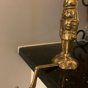 Puede incluir: Una figura de cascanueces de color latón con un gancho para colgar. La figura está detallada con un sombrero, uniforme y rasgos faciales. El gancho se extiende desde la base, diseñado para colgar en una estantería o repisa.