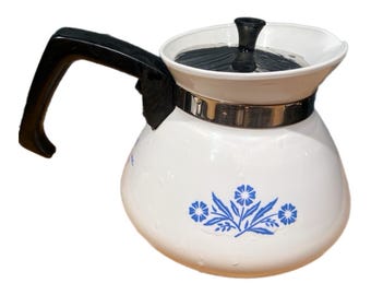 Op de afbeelding: Een witte keramische theepot met een blauw bloemmotief aan de voorzijde. De theepot heeft een zwart handvat, een zwart deksel met een knop en een zilverkleurige metalen band rond het bovenste gedeelte van de behuizing.