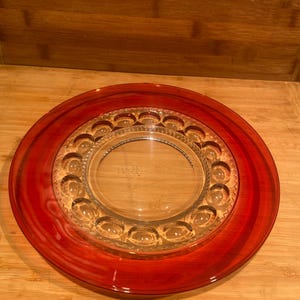 May include: A round glass plate featuring a wide, translucent red border and a clear centre. The inner section is decorated with a ring of raised, oval-shaped indentations and a delicate, textured geometric pattern surrounding the smooth middle.