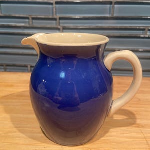 May include: A small ceramic pitcher featuring a deep cobalt blue glazed body with a contrasting cream-colored interior, rim, and handle. The vessel has a rounded, bulbous shape with a distinct pouring spout on one side and a curved handle.