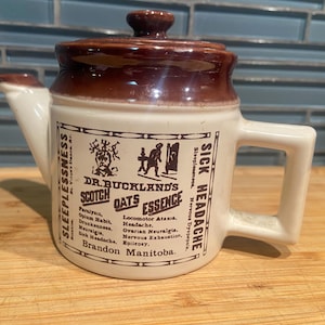 May include: A cream-coloured ceramic teapot with a brown lid and spout base. The body features black printed text reading Dr. Buckland's Scotch Oats Essence, Brandon Manitoba, and a list of ailments including Sleeplessness and Sick Headache.