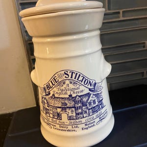 Könnte beinhalten: Cremefarbener Keramiktopf mit Deckel, mit einer blauen Grafik eines Gebäudes und dem Text "BLUE STILTON Traditional English Cheese". Der Topf hat eine zylindrische Form mit breiterem Oberteil und kleinem Griff.