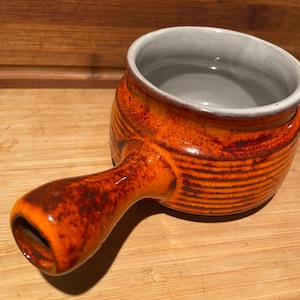 Könnte beinhalten: Eine kleine orangefarbene Suppenschüssel aus Keramik mit grauem Innenraum und einem Henkel. Die Schüssel hat ein strukturiertes, gestreiftes Design.