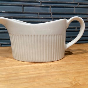 May include: A white ceramic gravy boat with a handle and a spout. The boat has vertical ribbed detailing on the sides. It is sitting on a wooden surface, with a blue tile background.