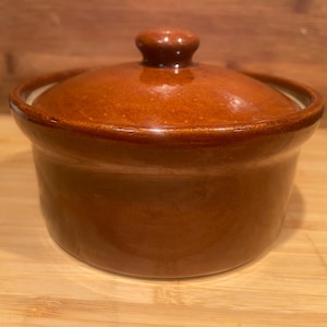 May include: A round, glazed ceramic casserole dish with a matching lid. The exterior features a glossy, speckled brown finish. The lid has a small, rounded knob handle at the centre. The interior of the dish is a light, neutral cream colour.