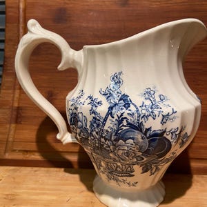 May include: A white ceramic pitcher with a blue floral pattern. The pitcher has a fluted body and a decorative handle.