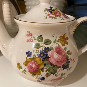 May include: A white ceramic teapot with a lid, decorated with a floral design of pink, yellow, and blue flowers. The teapot has a gold trim and a curved handle, ideal for serving tea.