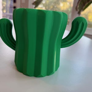 May include: A green cactus-shaped planter with a ribbed texture. The planter has two arms that extend out from the sides.