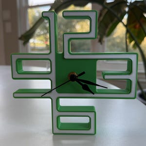 Puede incluir: Un reloj verde y blanco con un diseño único. La esfera del reloj está rodeada por un borde blanco y los números están formados por el marco verde y blanco.