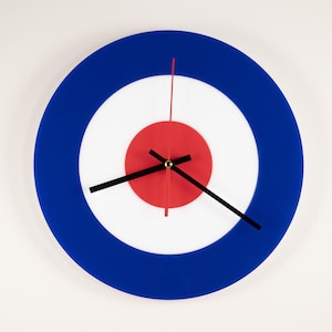 Puede incluir: Un reloj de pared redondo con un diseño de diana azul, blanco y rojo. La esfera del reloj presenta un anillo exterior azul, un anillo central blanco y un círculo central rojo. Las manecillas negras indican la hora. Un elemento decorativo.
