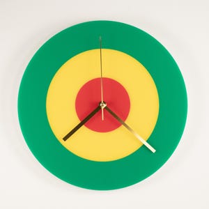 Puede incluir: Un reloj de pared redondo con diseño de diana. La esfera del reloj presenta círculos concéntricos en verde, amarillo y rojo. Las manecillas del reloj son doradas y el reloj está sobre un fondo blanco.