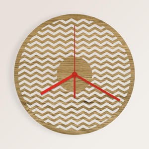 May include: A round wooden wall clock with a light brown finish. The clock face features a white zigzag pattern and red hands. The clock hands are red and the overall design is modern and minimalist.