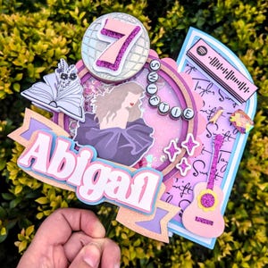 Peut inclure: Décoration de gâteau multicouche avec le nom "Abigail" en blanc et rose, un portrait, une guitare et le chiffre 7. La décoration a une palette de couleurs rose et violet avec des accents de paillettes. Le design comprend un code Spotify et le mot "Swiftie".