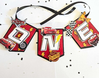 Puede incluir: Una guirnalda roja y negra con la palabra "ONE" en letras blancas. Presenta personajes de la película Cars, incluyendo a Rayo McQueen. La guirnalda está decorada con rayos y una cinta negra, ideal para un primer cumpleaños.