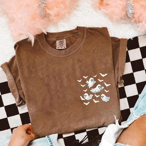 May include: Brown t-shirt with a pocket design featuring white ghosts and bats. The shirt has a crew neck and short sleeves. The tag reads "Comfort Colors". The shirt is a relaxed fit.
