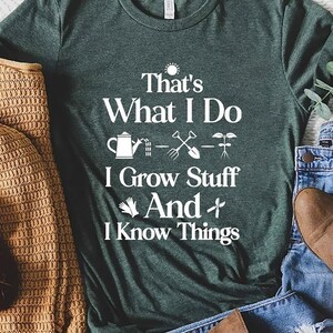 May include: A dark green t-shirt with the text "That's What I Do, I Grow Stuff And I Know Things" in white. The shirt is paired with blue jeans, brown boots, and a tan sweater.