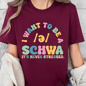 May include: Maroon crew neck t-shirt with the text "I WANT TO BE A SCHWA, IT'S NEVER STRESSED" in colorful, retro-style lettering. The shirt is a solid maroon color.